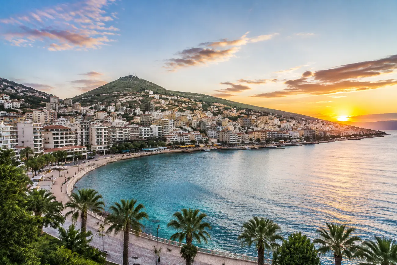 Sarandë şehri ve limanı gün batımında, arkada tepe ve palmiyeli sahil bulvarı